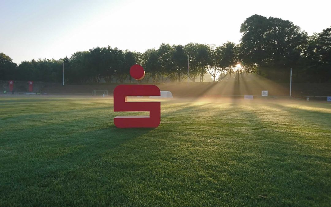Rotes Sparkassen-S auf grüner Wiese im Stadion Gladbeck im Morgendunst und Gegenlicht, mit Sonne, die im Hintergrund durch die Bäume bricht
