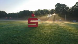 Rotes Sparkassen-S auf grüner Wiese im Stadion Gladbeck im Morgendunst und Gegenlicht, mit Sonne, die im Hintergrund durch die Bäume bricht